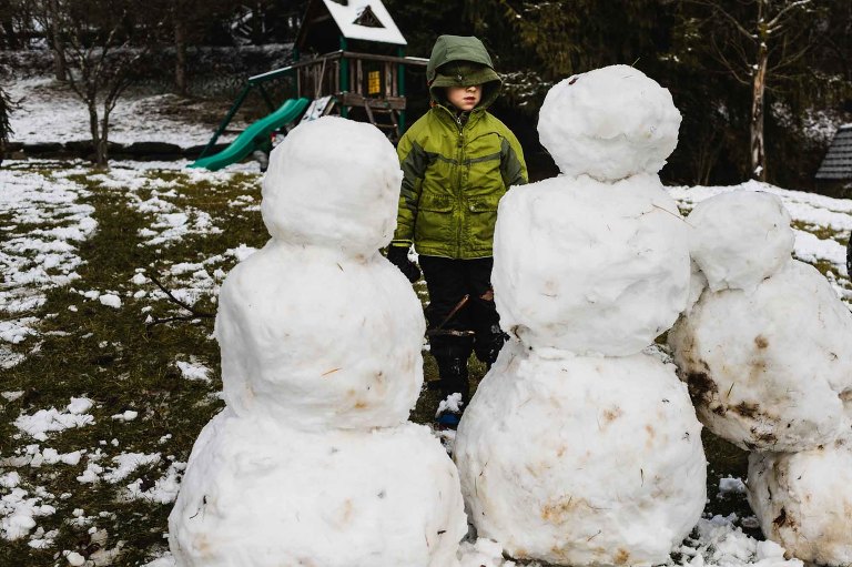 boy bundled up for winter weather, eyes hidden by his coat hood, standing in front of and looking at three snowmen. 