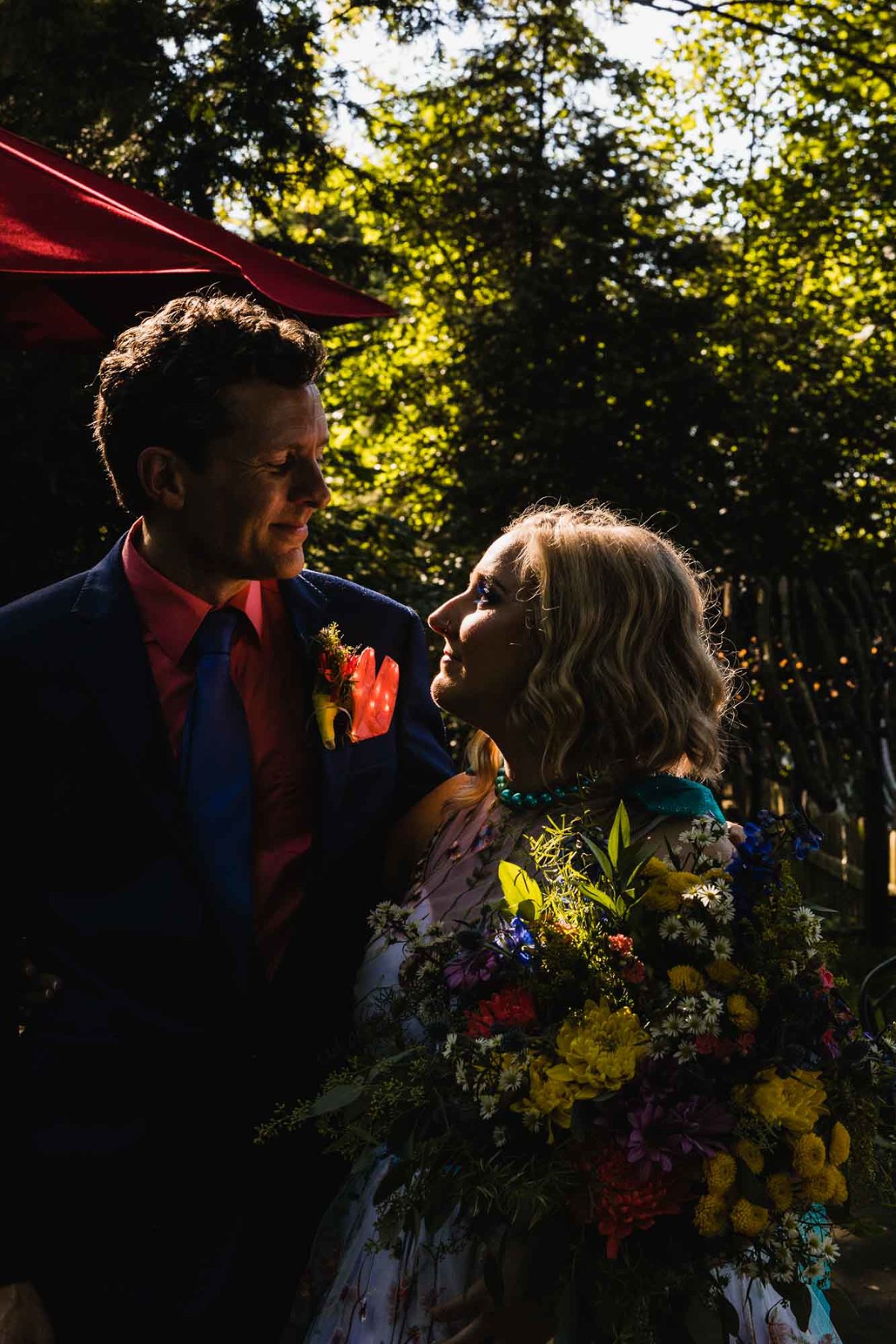 bride and groom look into each others' eyes, highlighted by the sun in front of green trees