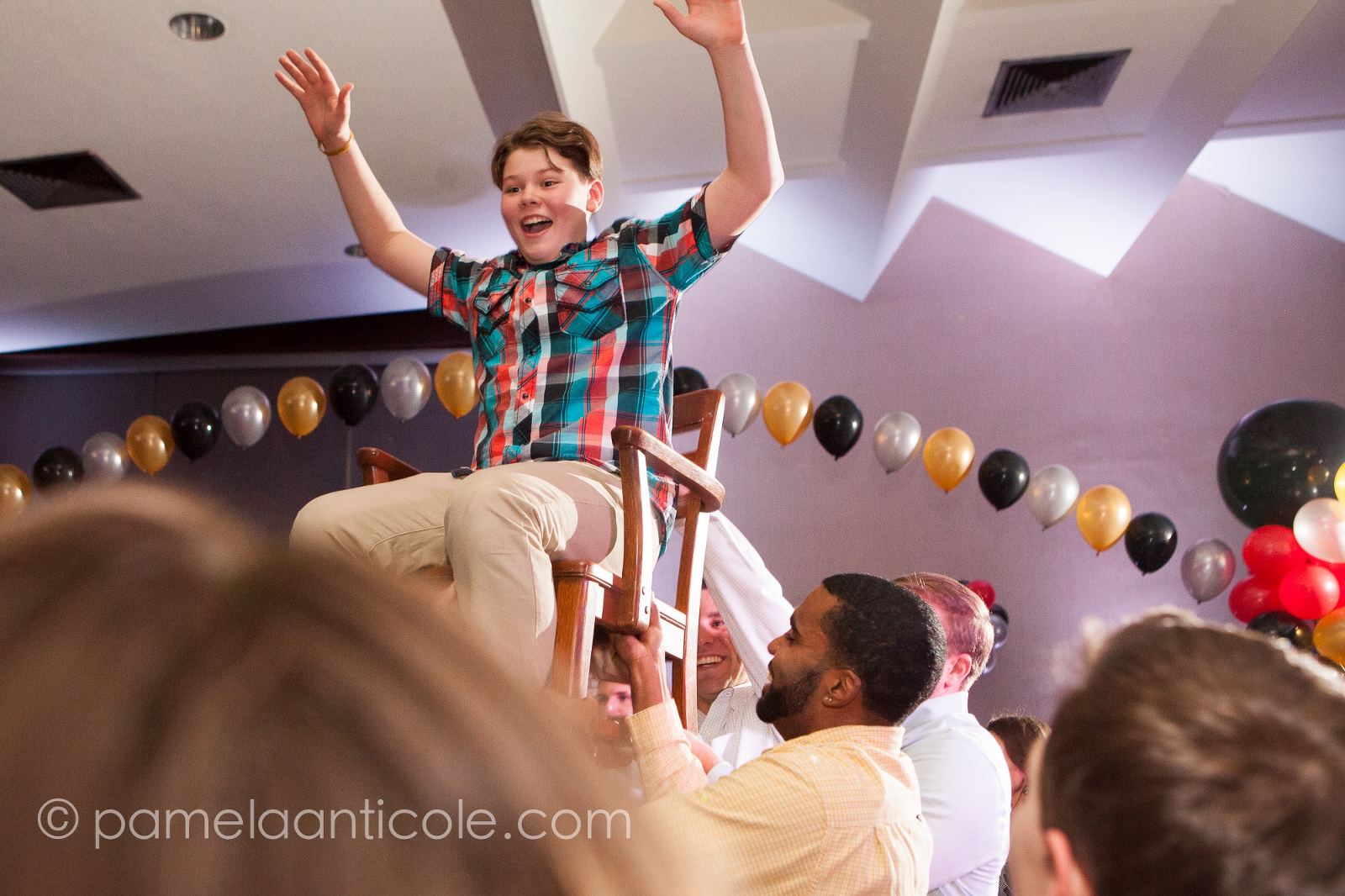 mitzvah boy goes up on a chair at a party, surrounded by friends and family and balloons