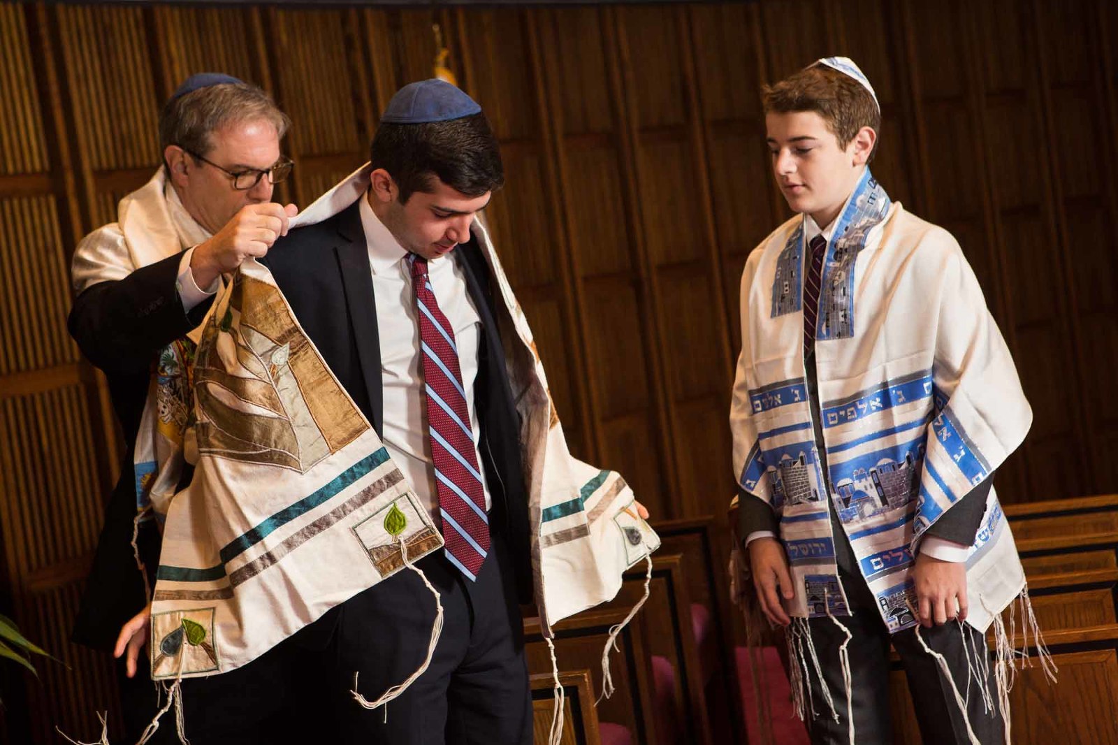 dad helps his bar mitzvah son put on his tallit before his mitzvah service, as big brother looks on. 