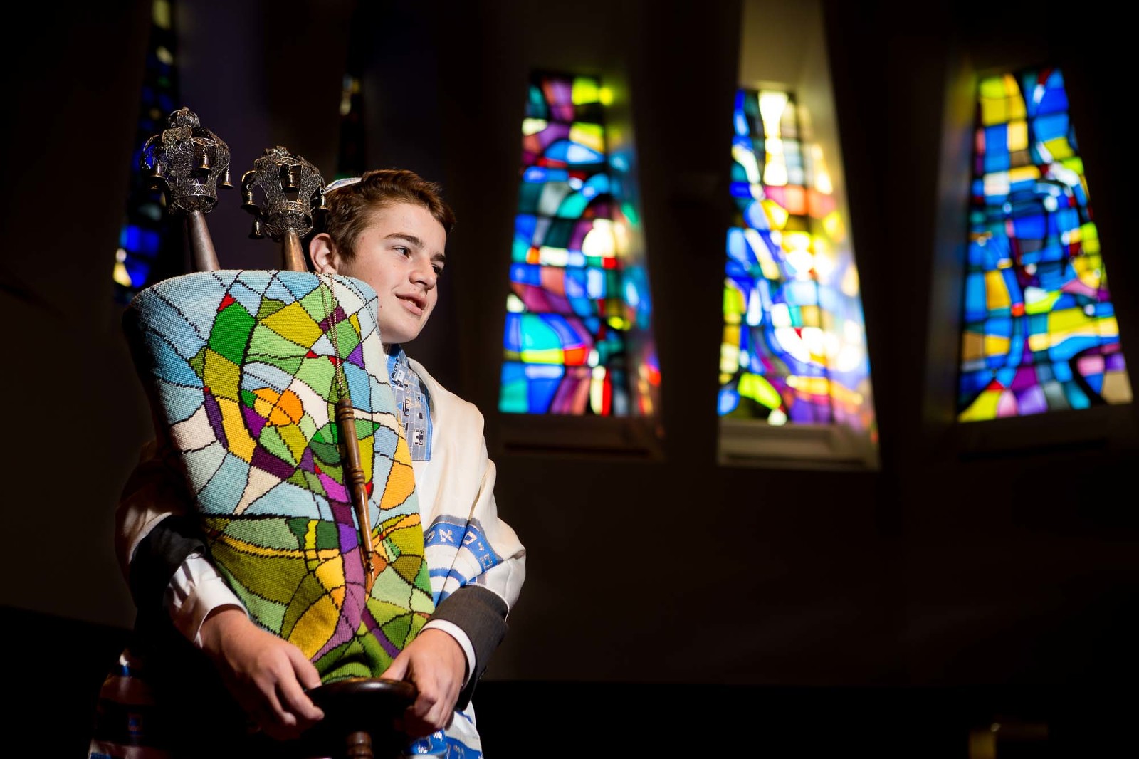 bar mitzvah boy stands holding the torah in front of some stained glass windows.