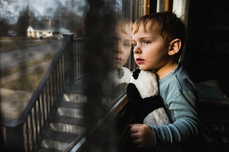 boy sits at the window with smeared lipstick and panda bear stuffed animal, looking out the window