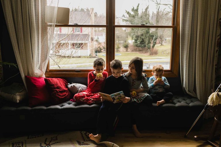 kids sit together in a window seat, reading and eating apples