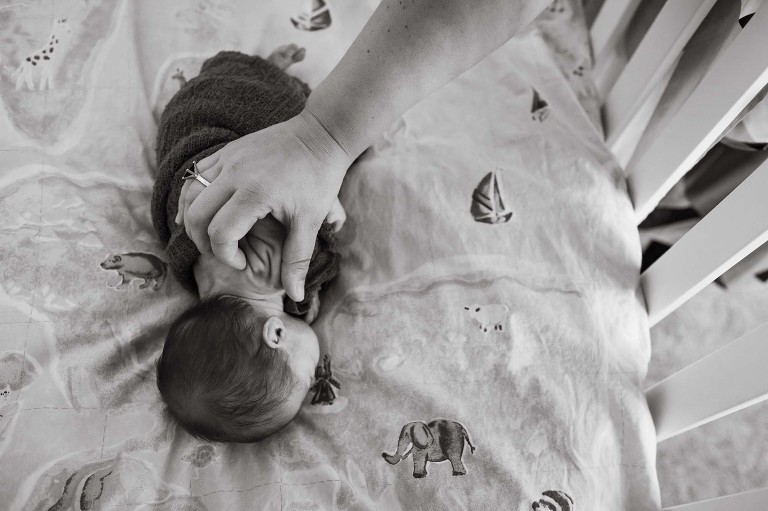 black and white photo of baby girl lying in crib with mom petting wrinkles on her back