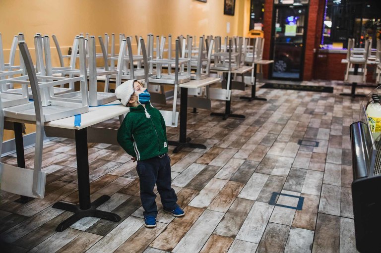 tired little boy wearing mask, leaning against empty tables in a restaurant in pittsburgh