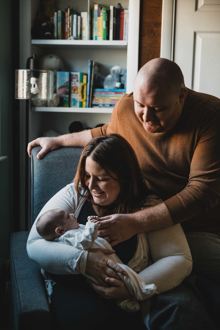 mom cradles baby boy in a chair, while dad props himself on the armchair and smiles down at newborn baby. all photographed in baby's nursery in front of books, with beautiful window light
