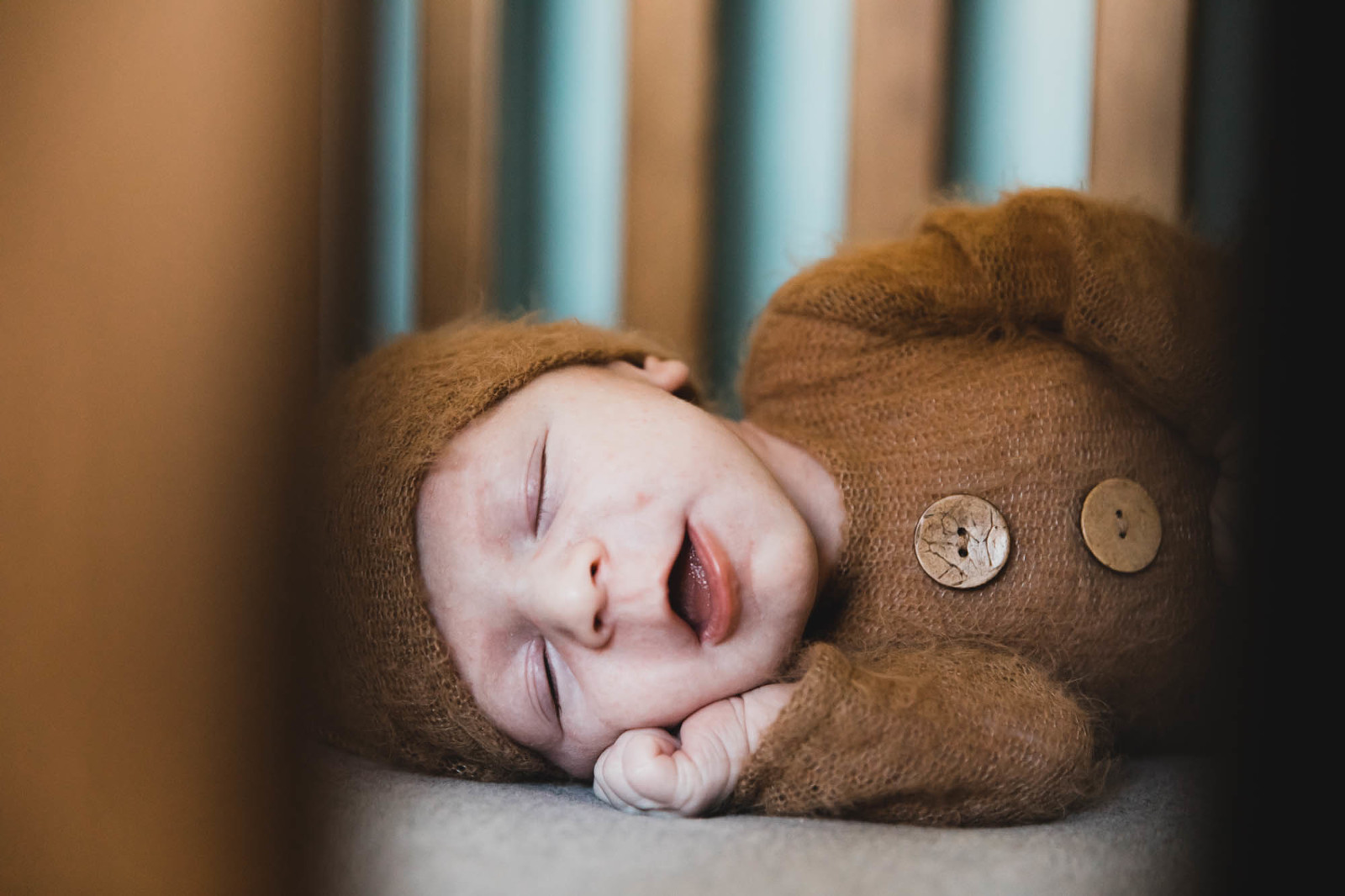 sleeping baby smiles in crib, bundled up in adorable brown onesie with wood buttons and hat