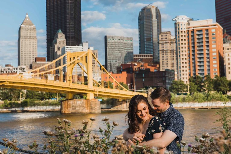 man and woman snuggle together in front of the city of pittsburgh, lit beautifully by golden hour