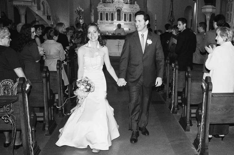 bride and groom exit church holding hands and smiling, in black and white