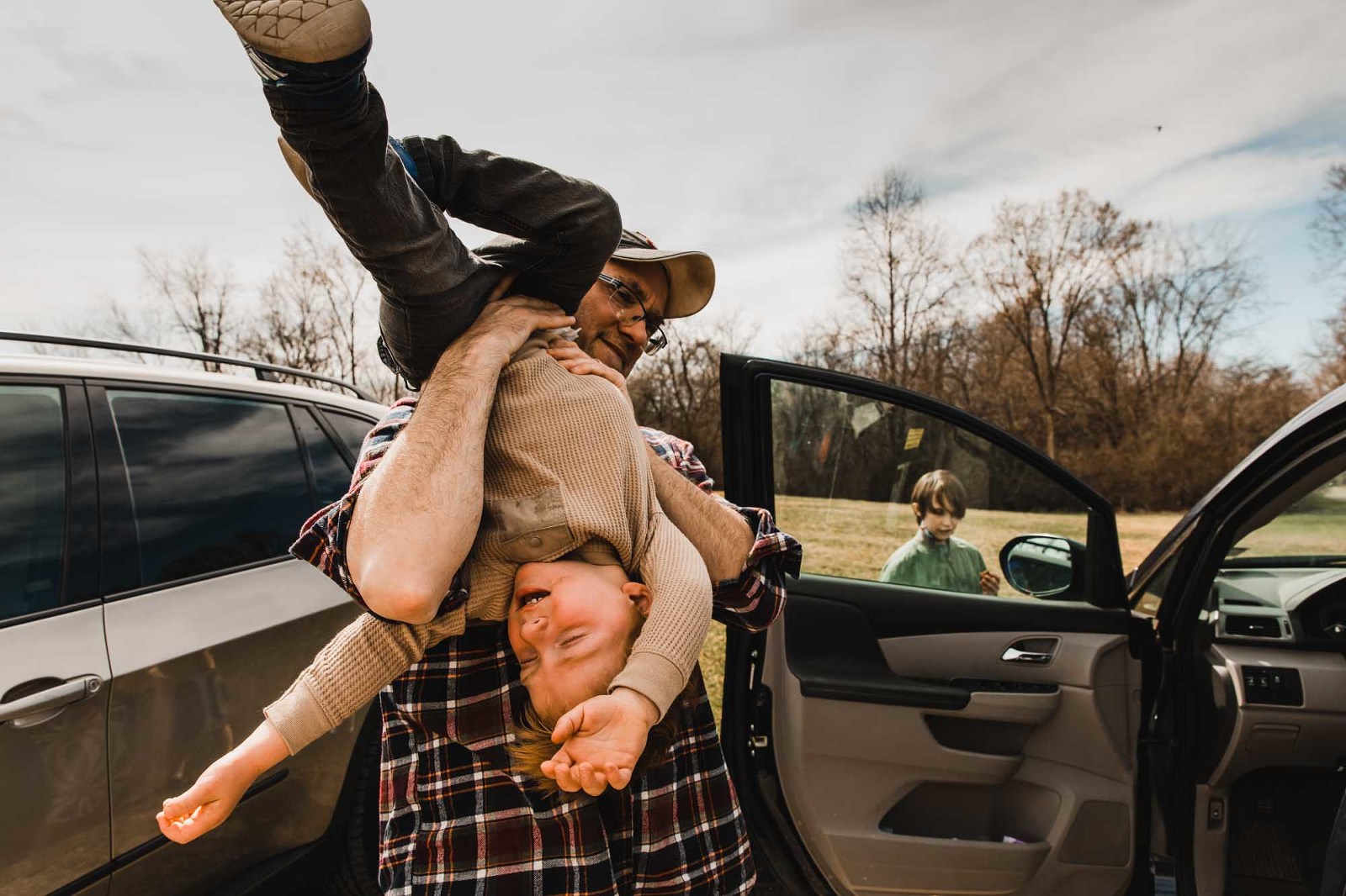 toddler throwing a tantrum, being held upside down by his dad, standing next to the car in a park