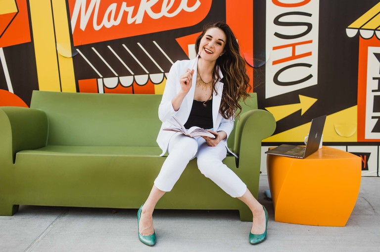 woman sits on green couch surrounded by retro themed colorful mural wall, drawing in a sketchbook and acting silly