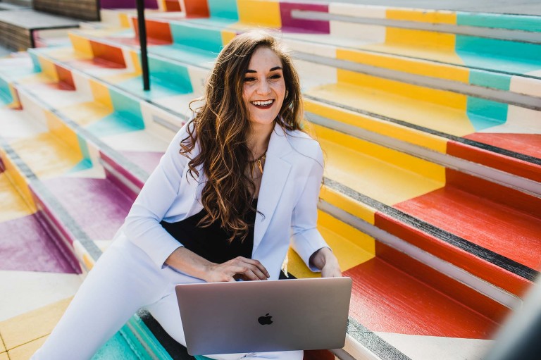 woman in white pantsuit sits at her laptop on colorful steps, smiling and looking away