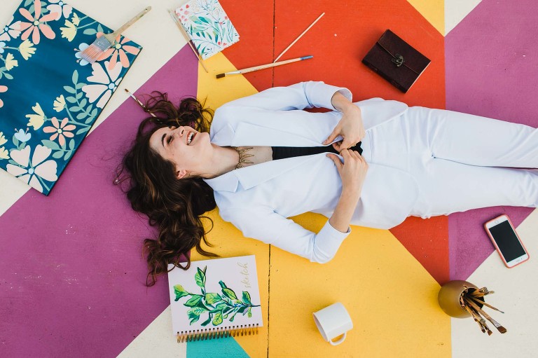 woman lies on colorful painted ground surrounded by paintbrushes, canvas, and sketchpad