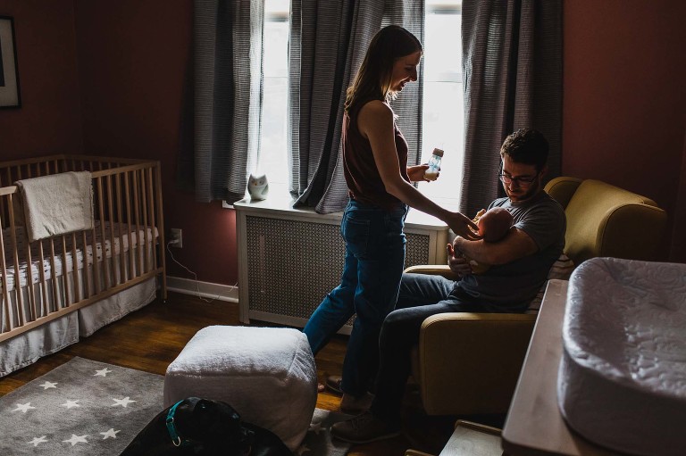 moody photo of a baby's nursery, with dad holding baby in a chair and mom reaching out to touch baby's head