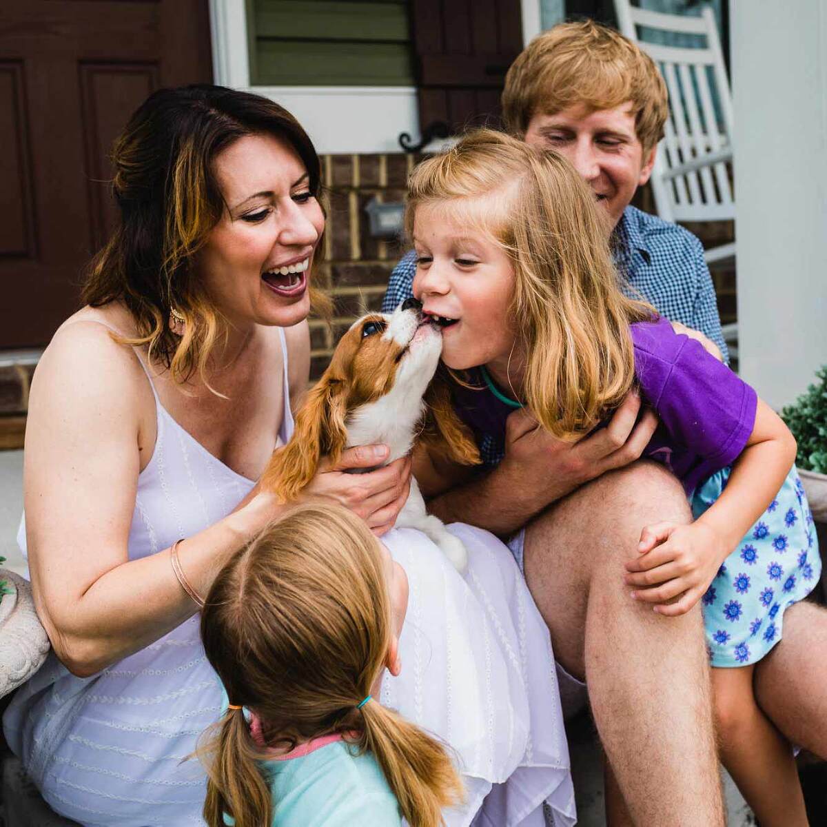 candid picture of a family sitting on their front porch, with a little girl giving a puppy kisses and everyone laughing