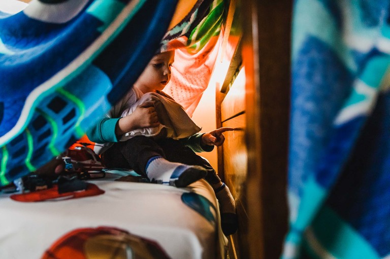 color photo of little boy playing underneath a blanket fort, in a pensive moment drawing his finger across the footer of his bed