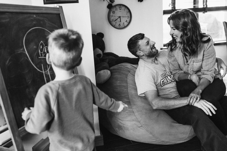black and white picture of candid moment between mom and dad as a young boy draws on a chalkboard