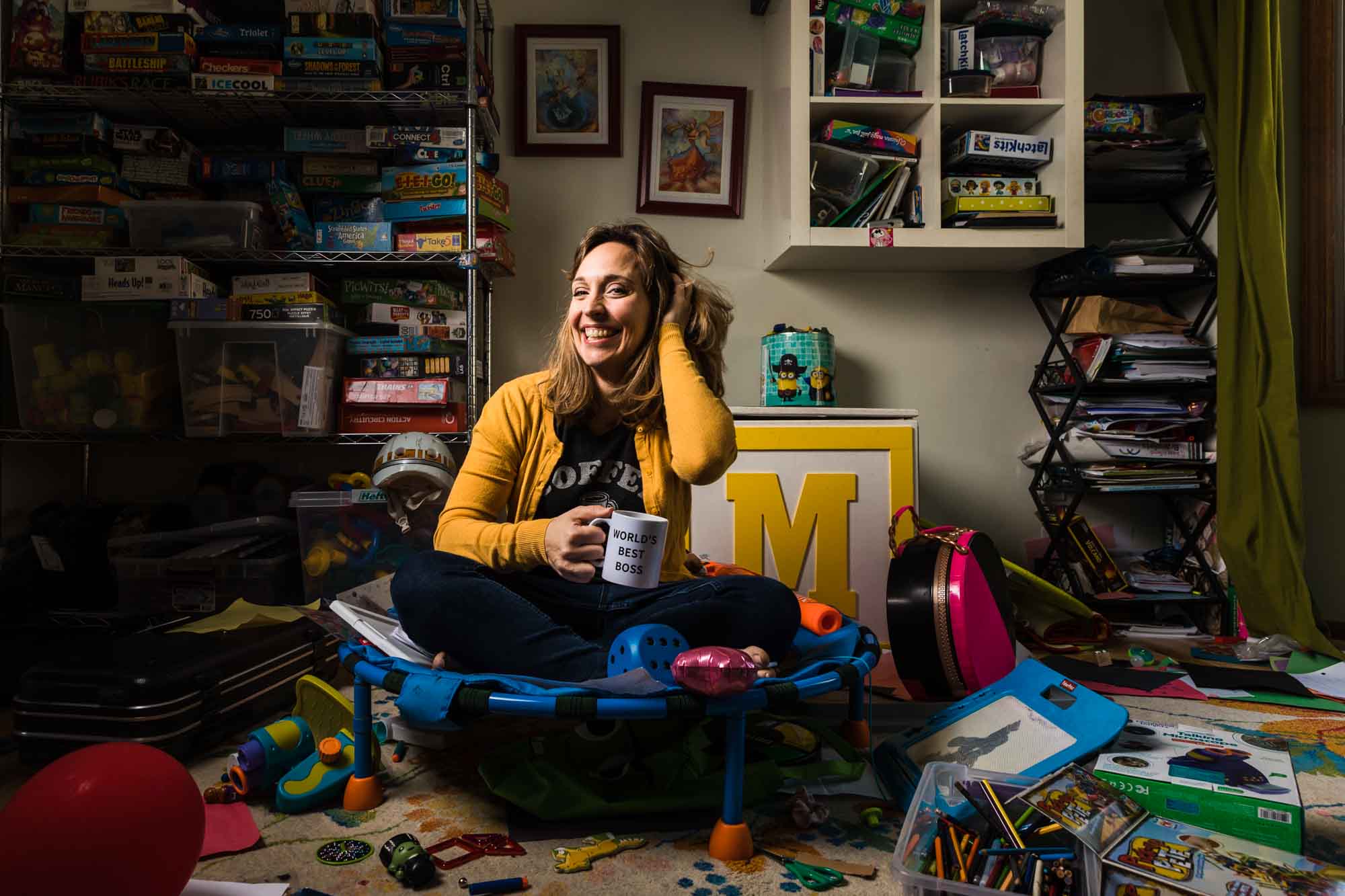 environmental portrait of pittsburgh photographer mom in yellow sweater sitting on a trampoline in a messy playroom, holding a mug that says world's best boss