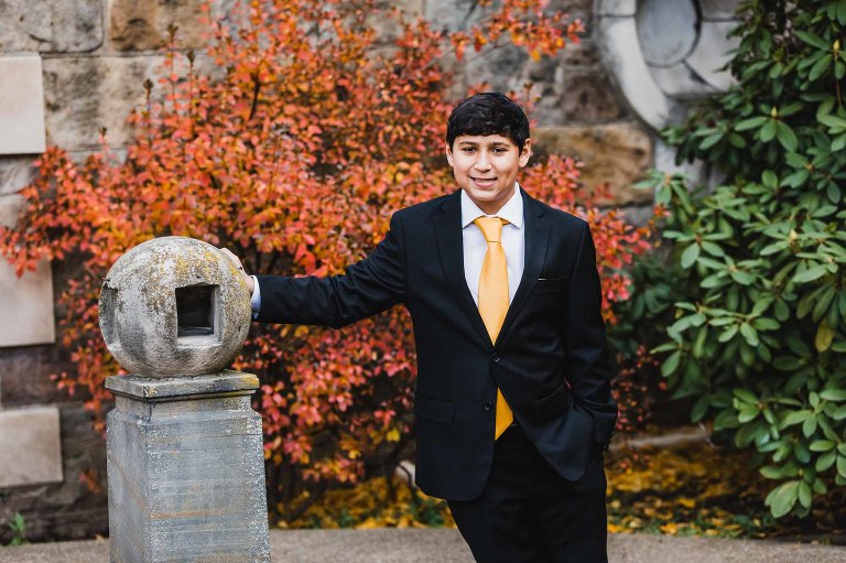 portrait of young boy in a black and gold suit, leaning against a pillar in front of an orange tree