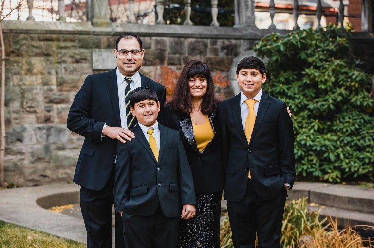 family with mom, dad, and two young boys wearing suits and ties in black and gold, standing together in a city garden scene