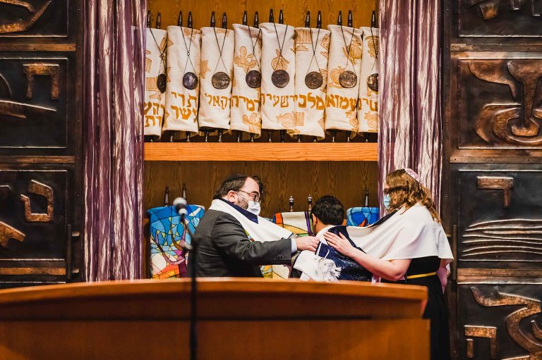 young boy stands in front of the ark full of torahs, at his bar mitzvah service, while the rabbi lays his tallit on the bar mitzvah boy's shoulders