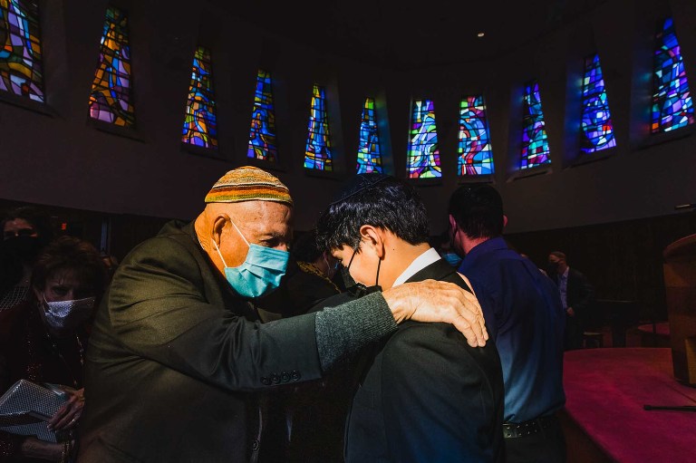 older Jewish man puts his hand on his grandson's shoulder and prays for him, in front of some stained glass windows in the synagogue
