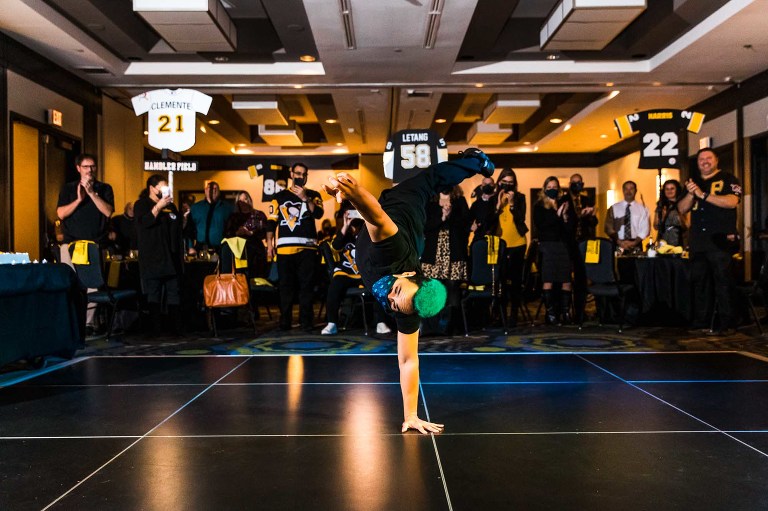 breakdancing man with green hair, standing on his hand on a dance floor, surrounded by black and gold table decor in a hotel ballroom area