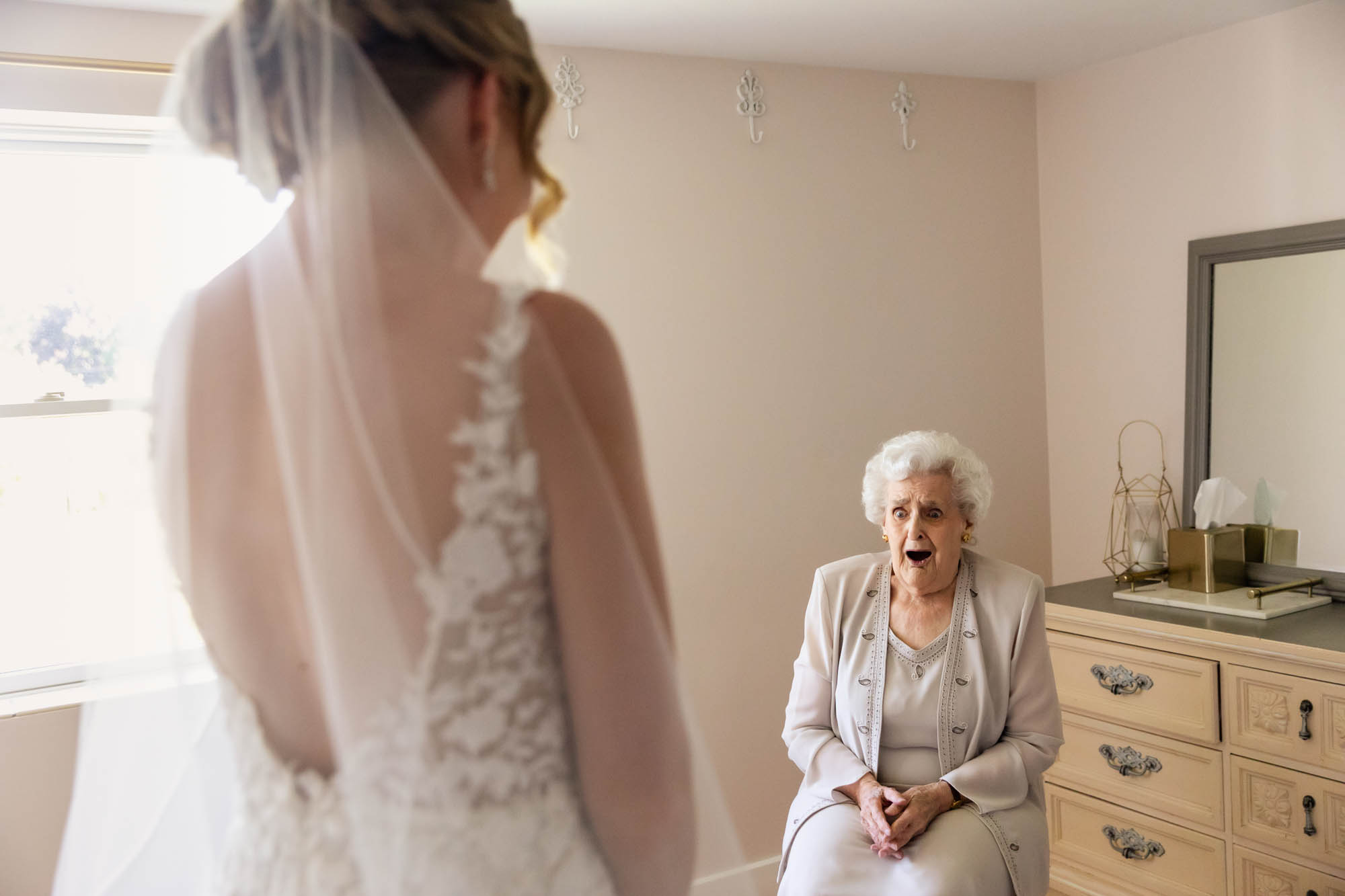 elderly woman dressed formally sitting in a chair in a room, with her hands crossed in her lap and a shocked happy expression on her face- looking at a bride standing across the room on her wedding day