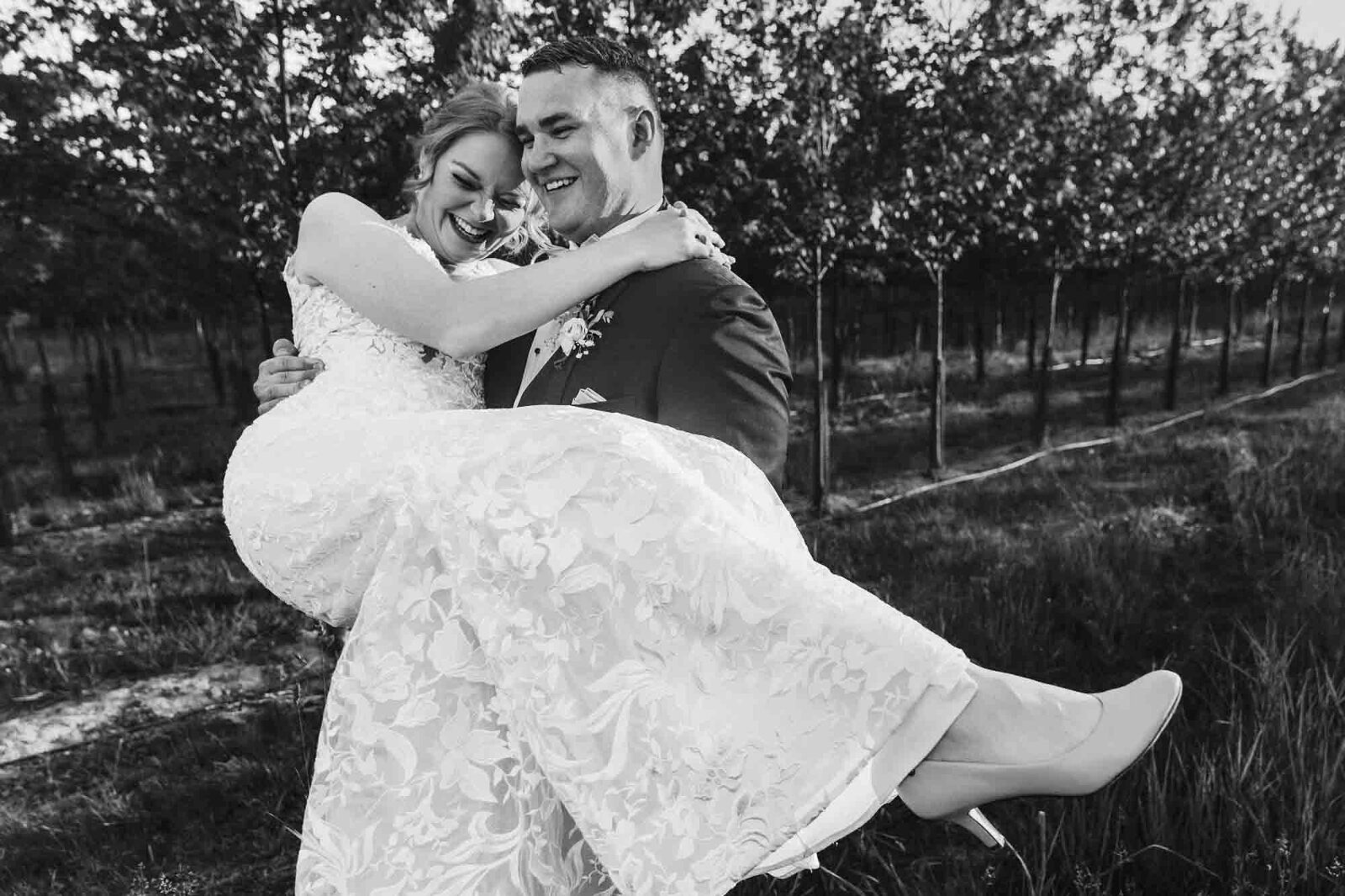 man carries woman in black and white, apparently wearing a wedding dress, through an orderly field at sunset. black and white photo taken by documentary wedding photographer in pittsburgh pa
