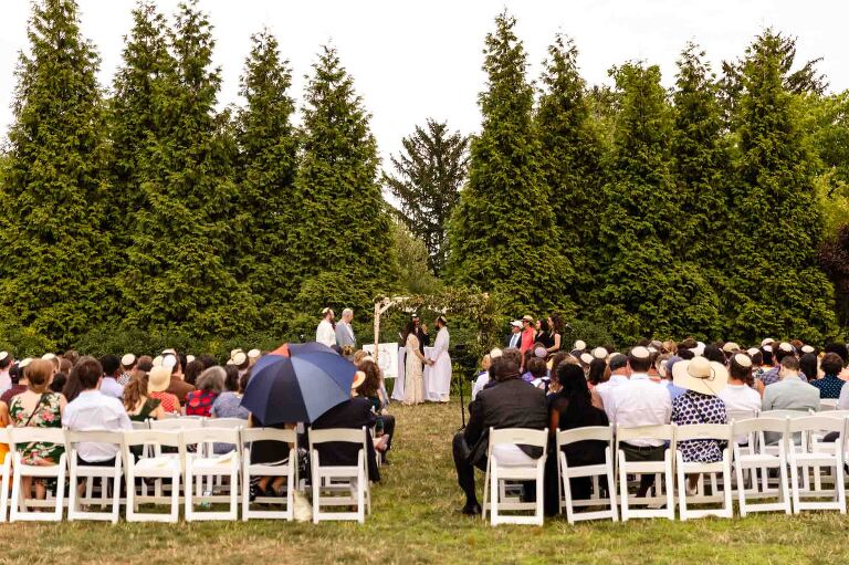 couple gets married under a chuppah in front of a natural setting of tall trees, with folding chairs set out on a lawn