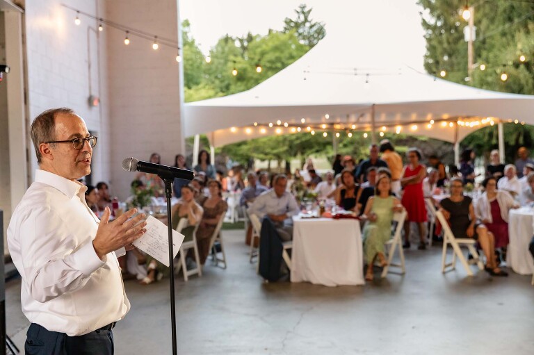 man gives speech to wedding guests at an outdoor wedding venue, aspinwall riverfront park. you can see trees and a tent in the background.
