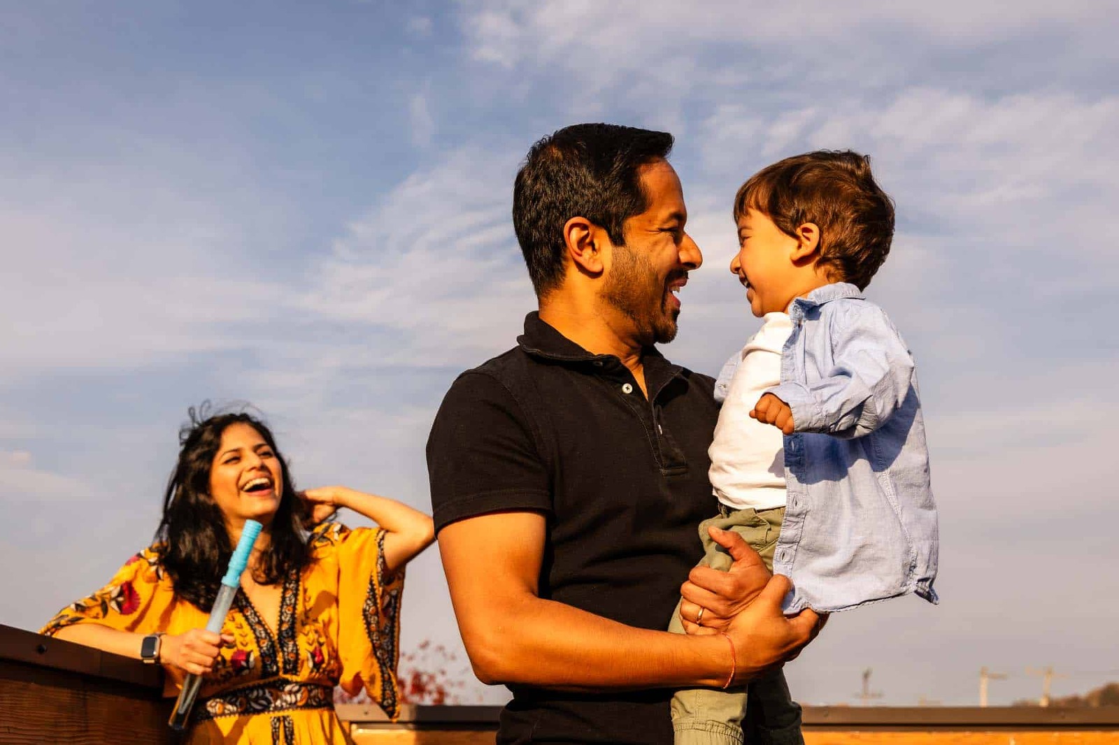 man and boy laugh together while woman watches on laughing in the background. surrounded by blue sky on a sunny day. picture taken by professional photographer in pittsburgh, pamela anticole