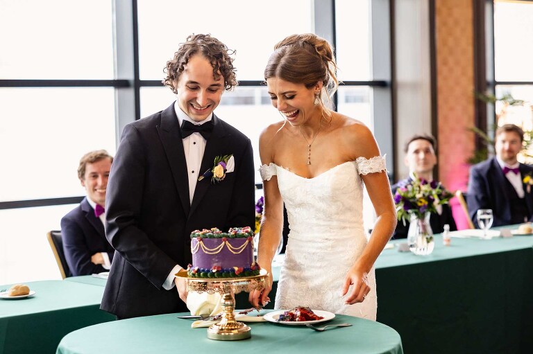 bride and groom cut into their wedding cake laughing in duquesne power center ballroom overlooking windows over the city of pittsburgh