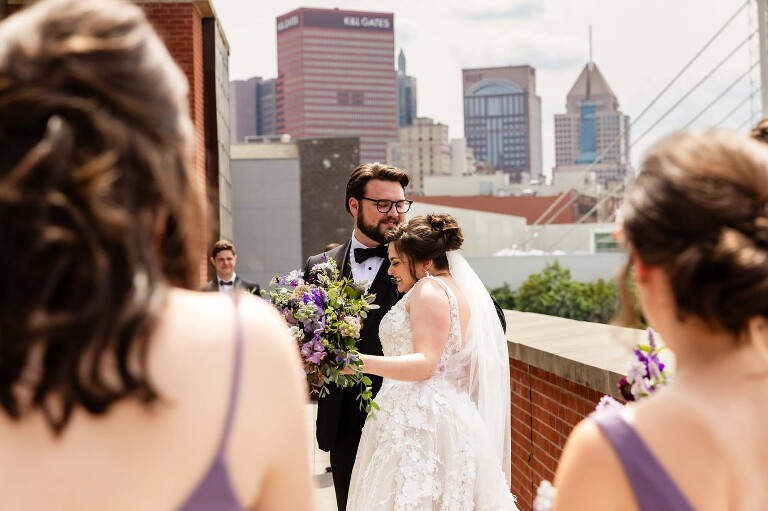 bride and groom snuggle together surrounded by their best friends and bridal party. taken on the balcony at the heinz history center wedding venue, outside the mueller center. candid pictures taken by pittsburgh wedding photographer pamela anticole