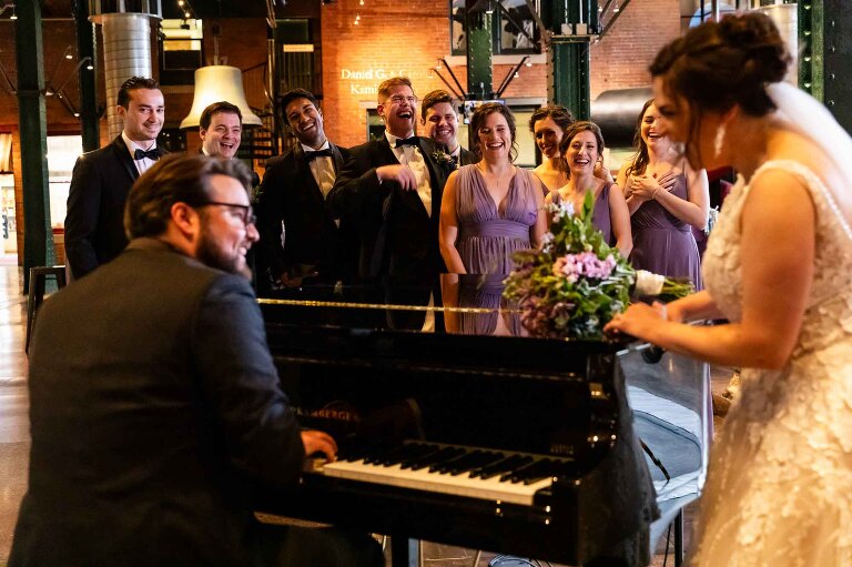 bride and groom sing together on the piano in the grand hall at their heinz history center wedding. bridal party looks on in the background.