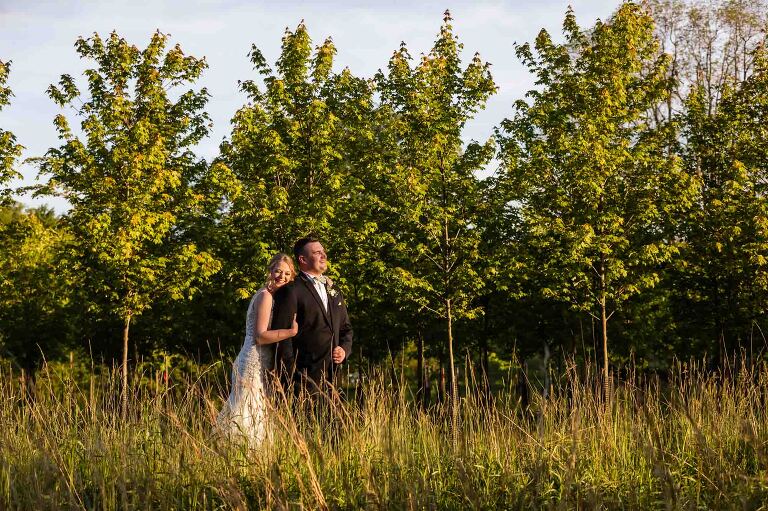 bride and groom snuggle together in front of a row of trees at sunset