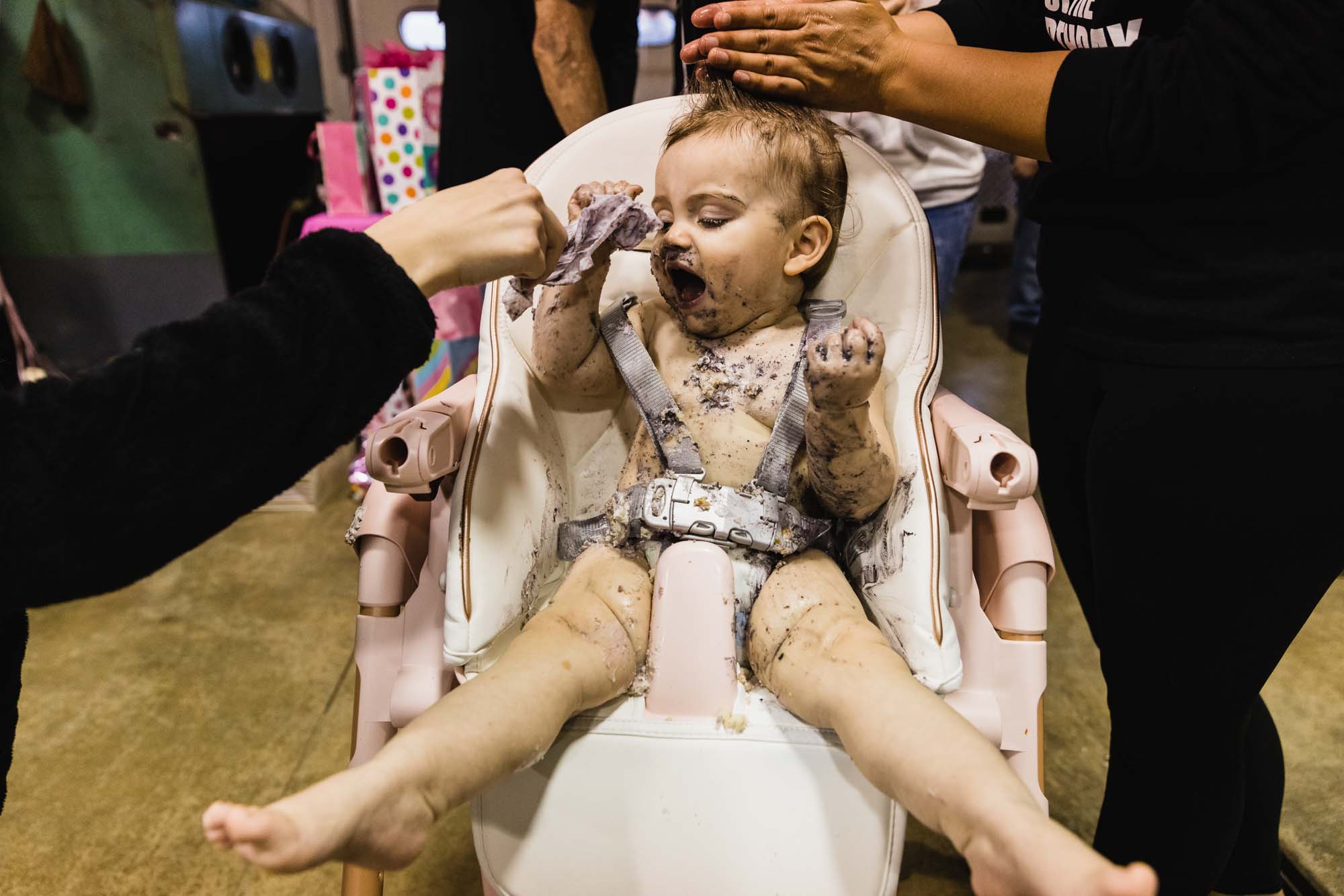 photo of little girl in her diaper in her high chair covered in cake being wiped down by her parents